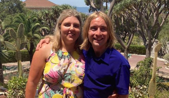 Warren and Elin stand together in a tropical garden setting, both smiling warmly. Elin has blonde hair and is wearing a sleeveless dress with a vibrant floral pattern in shades of pink, yellow, and green. Warren is wearing a deep blue shirt. The couple are happy and relaxed in the bright, natural light.